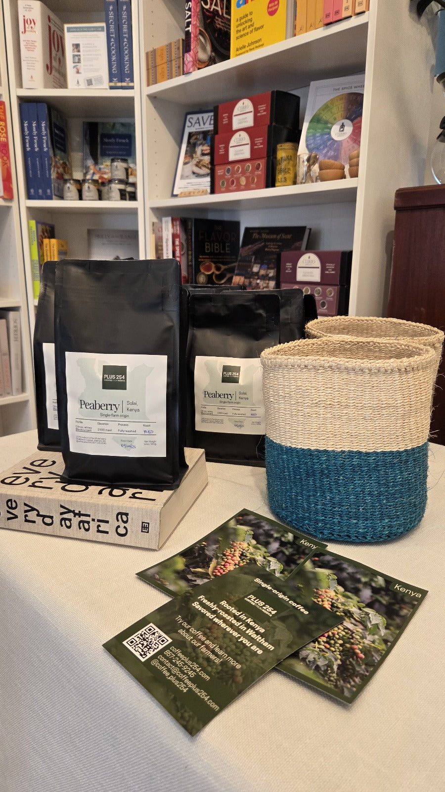 Two black bags of peaberry coffee with postcards from Coffee Plus 254 on a table next to business cards and a woven basket.