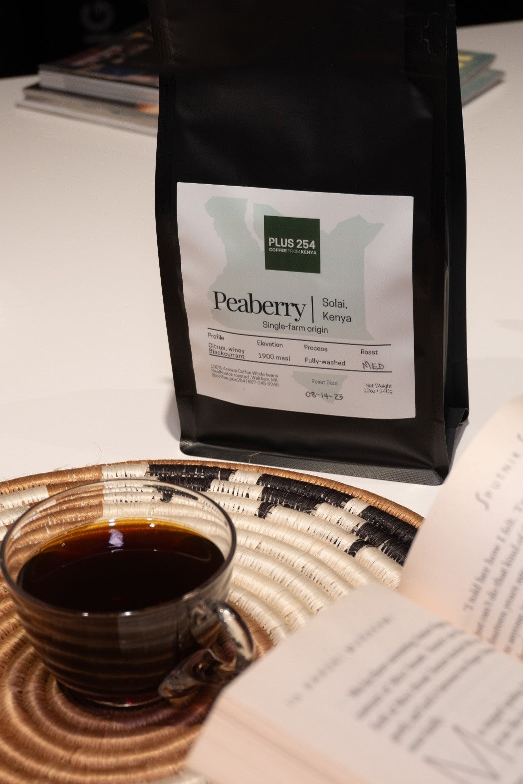 Bag of Peaberry coffee and a cup of coffee on a textured surface with an open book.