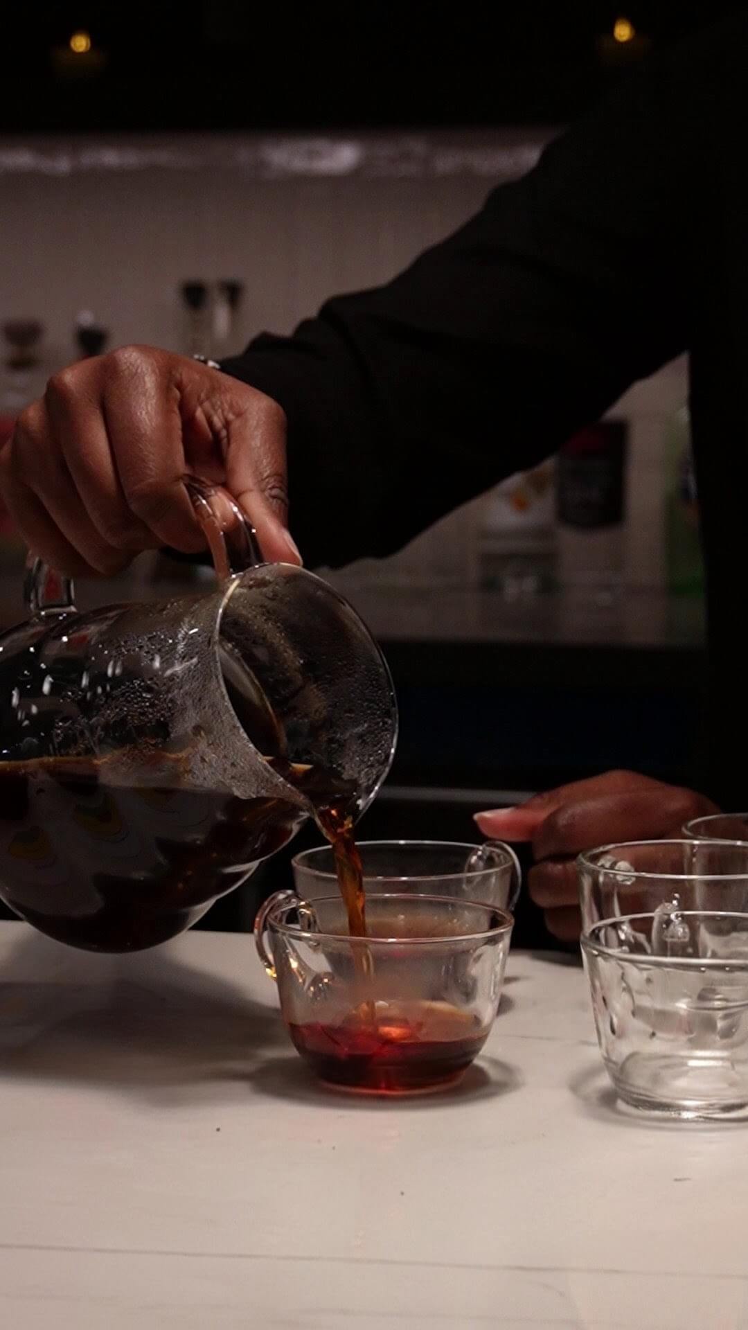 A person pouring dark coffee from a glass carafe into a small glass cup on a tabletop, with additional empty glasses nearby in a dim indoor setting.