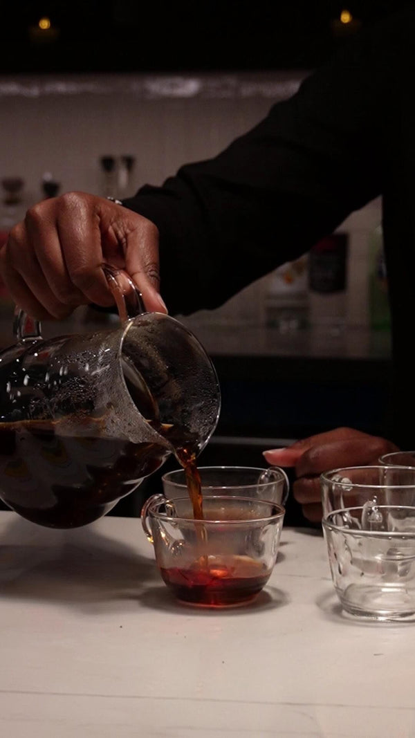 A person pouring dark coffee from a glass carafe into a small glass cup on a tabletop, with additional empty glasses nearby in a dim indoor setting.