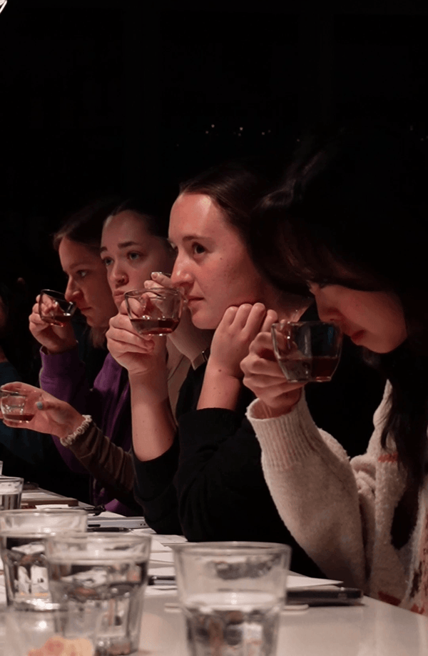 Four women seated at a table, each holding a small clear glass cup, tasting coffee and looking forward in a dimly lit indoor setting.